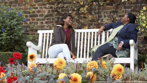 Two visitors sitting on a white bench laughing in the garden at Fenton House and Garden, with a bed of dahlias visible in the foreground and a brick wall with climbing plants behind the visitors.